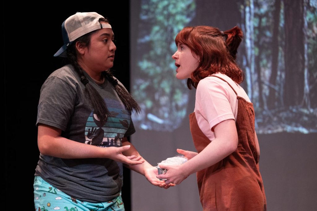 Two actors talking in front of a screen showing trees.